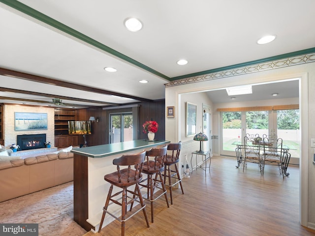 kitchen with a brick fireplace, light wood-type flooring, beamed ceiling, kitchen peninsula, and a kitchen bar