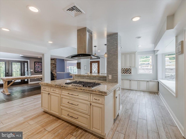 kitchen with island range hood, cream cabinets, stainless steel gas stovetop, light stone countertops, and light hardwood / wood-style floors