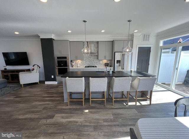 kitchen with appliances with stainless steel finishes, gray cabinets, wall chimney range hood, an island with sink, and a kitchen bar