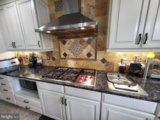 kitchen with decorative backsplash, dark stone counters, wall chimney range hood, stainless steel gas stovetop, and white cabinetry