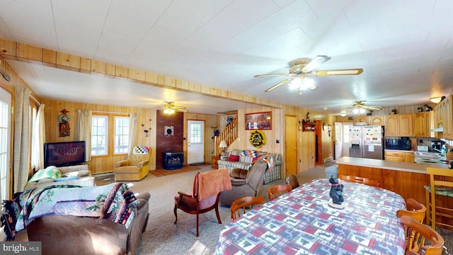 dining space with ceiling fan, light colored carpet, wood walls, stairs, and a brick fireplace