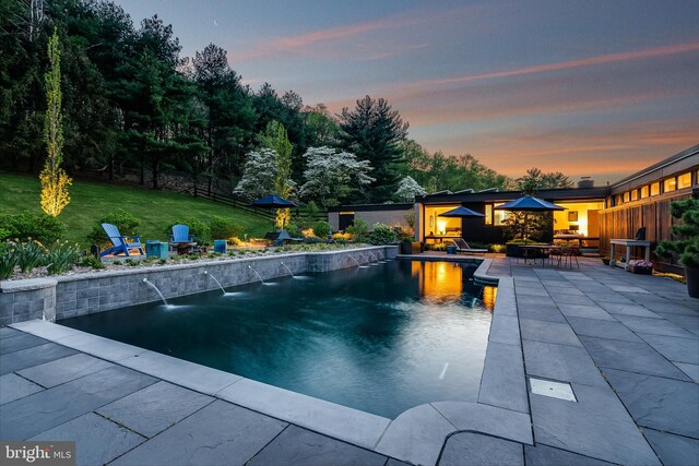 pool at dusk featuring pool water feature, a lawn, and a patio area