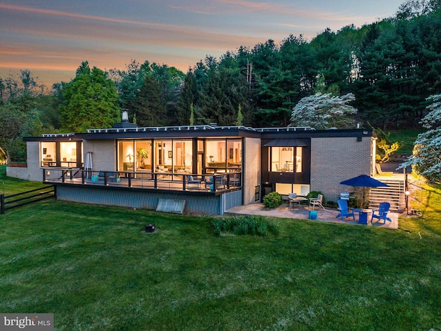 back house at dusk with a patio, a lawn, and a wooden deck