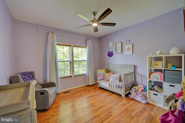 bedroom with a crib, baseboards, visible vents, ceiling fan, and wood finished floors