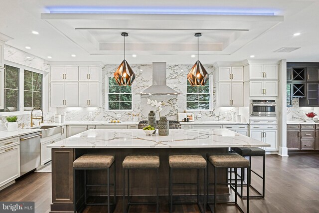 kitchen with hanging light fixtures, a center island, stainless steel appliances, wall chimney range hood, and a raised ceiling