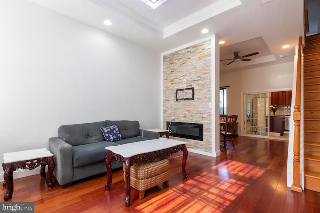 living room with dark hardwood / wood-style flooring, a tray ceiling, a stone fireplace, ceiling fan, and ornamental molding