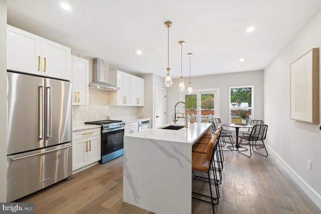 kitchen featuring white cabinets, stainless steel appliances, a center island with sink, sink, and wall chimney range hood