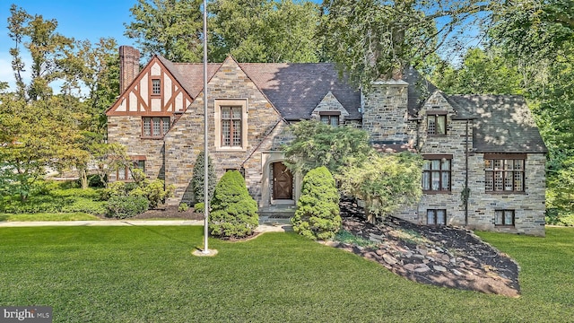 tudor-style house featuring stone siding, a chimney, a front lawn, and a high end roof