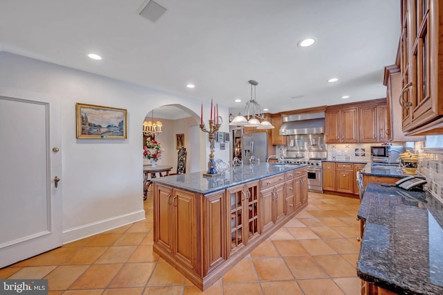 kitchen with arched walkways, a center island with sink, recessed lighting, appliances with stainless steel finishes, and wall chimney exhaust hood