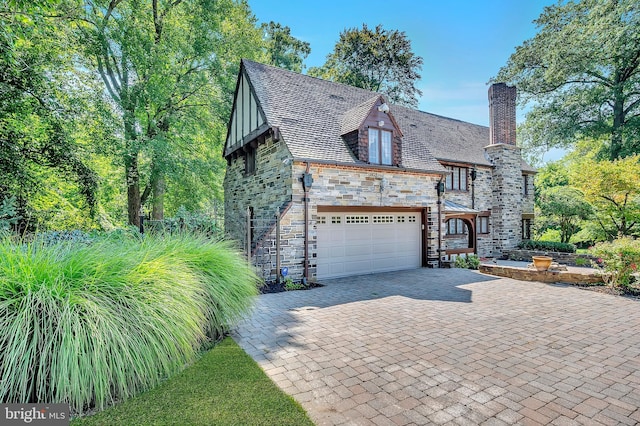 tudor-style house with a garage, stone siding, decorative driveway, a chimney, and a high end roof