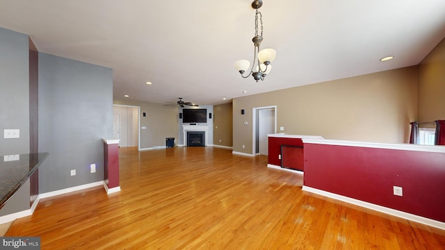 unfurnished living room featuring baseboards, a fireplace, light wood finished floors, and ceiling fan with notable chandelier