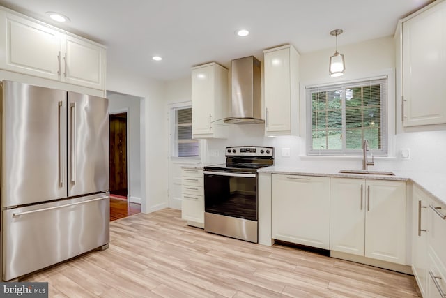 kitchen featuring stainless steel appliances, white cabinetry, wall chimney range hood, hanging light fixtures, and sink