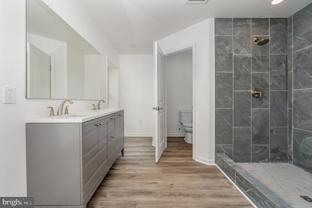 full bath featuring toilet, wood finished floors, tiled shower, double vanity, and baseboards