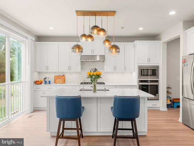 kitchen with light wood-type flooring, white cabinetry, an island with sink, hanging light fixtures, and appliances with stainless steel finishes