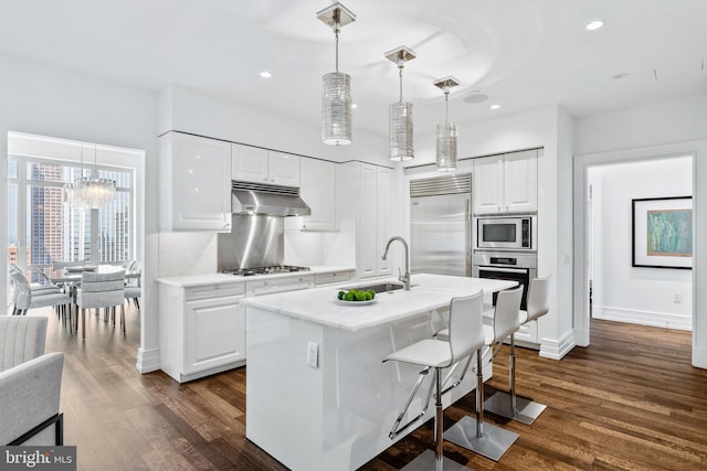 kitchen featuring sink, built in appliances, a center island with sink, white cabinetry, and hanging light fixtures