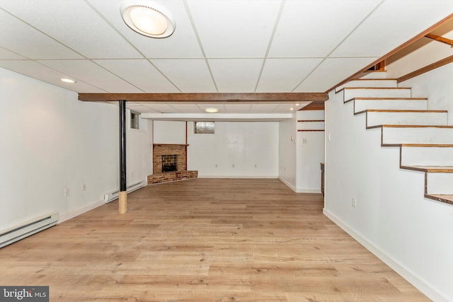 basement featuring a drop ceiling, light wood-type flooring, a brick fireplace, and a baseboard radiator