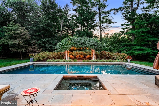 pool at dusk with a hot tub and a patio area