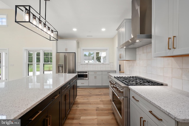 kitchen featuring light stone counters, light wood finished floors, high end appliances, tasteful backsplash, and wall chimney exhaust hood