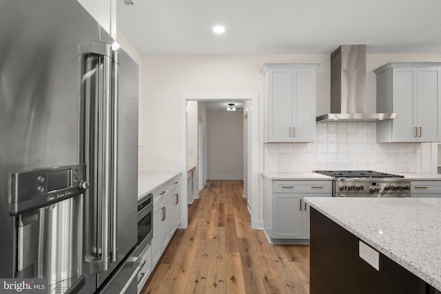 kitchen featuring light wood finished floors, stainless steel appliances, backsplash, light stone countertops, and wall chimney exhaust hood