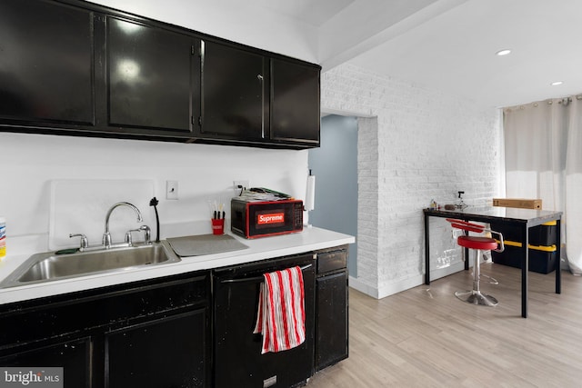 kitchen featuring dark cabinets, brick wall, a sink, light countertops, and light wood-type flooring