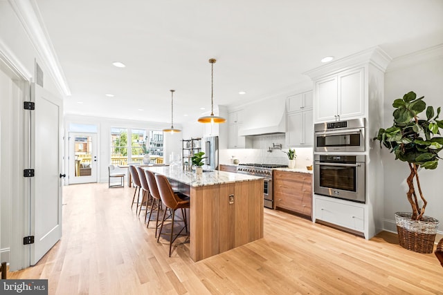 kitchen with a center island with sink, custom range hood, appliances with stainless steel finishes, white cabinetry, and light stone counters