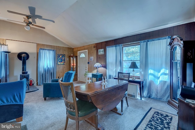 dining space with wooden walls, a wood stove, ceiling fan, lofted ceiling, and light colored carpet