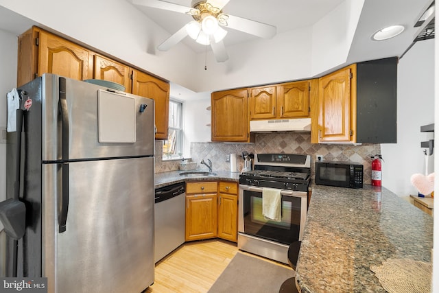 kitchen with stainless steel appliances, ceiling fan, light hardwood / wood-style flooring, dark stone counters, and sink