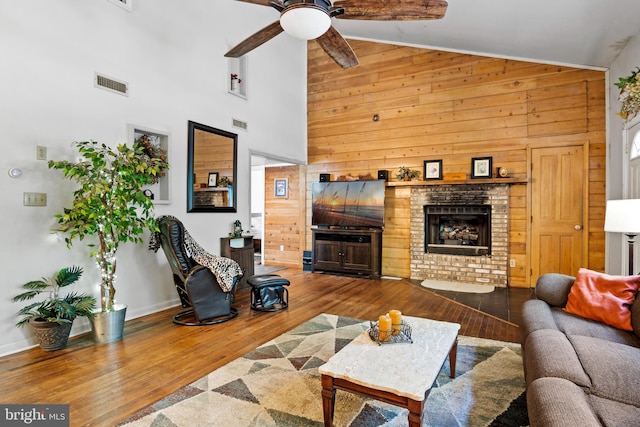 living room with wooden walls, a brick fireplace, hardwood / wood-style floors, ceiling fan, and high vaulted ceiling