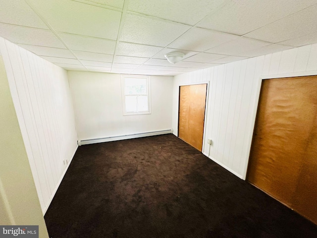 unfurnished bedroom with wood walls, baseboard heating, dark carpet, and a drop ceiling