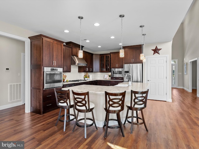 kitchen with appliances with stainless steel finishes, wall chimney range hood, an island with sink, dark wood-type flooring, and dark brown cabinetry