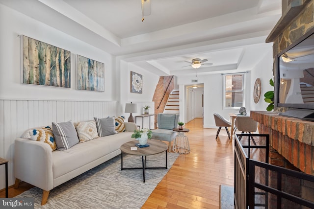 living area featuring light wood finished floors, a raised ceiling, wainscoting, ceiling fan, and stairs