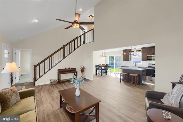 living area featuring stairway, light wood-style flooring, ceiling fan with notable chandelier, and high vaulted ceiling