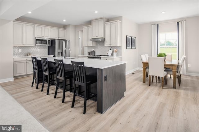 kitchen featuring backsplash, light hardwood / wood-style flooring, appliances with stainless steel finishes, a kitchen island with sink, and a breakfast bar