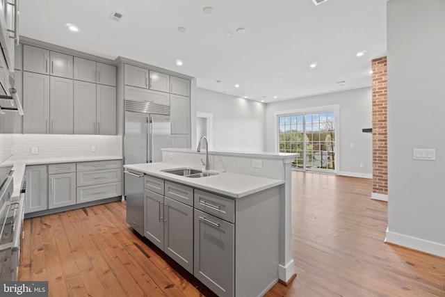 kitchen with gray cabinetry, appliances with stainless steel finishes, an island with sink, sink, and light wood-type flooring