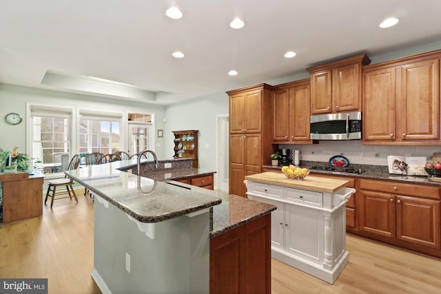 kitchen featuring light wood-type flooring, backsplash, an island with sink, and dark stone counters