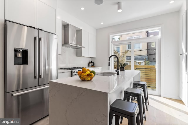 kitchen with wall chimney exhaust hood, an island with sink, stainless steel appliances, light stone counters, and white cabinets