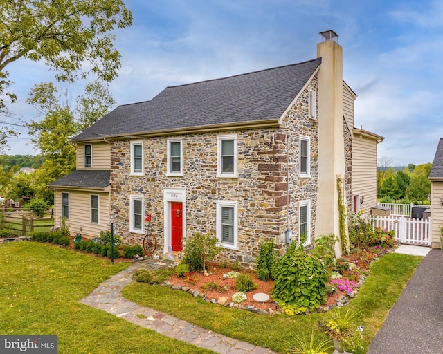 view of front of house with a front lawn, stone siding, fence, roof with shingles, and a chimney