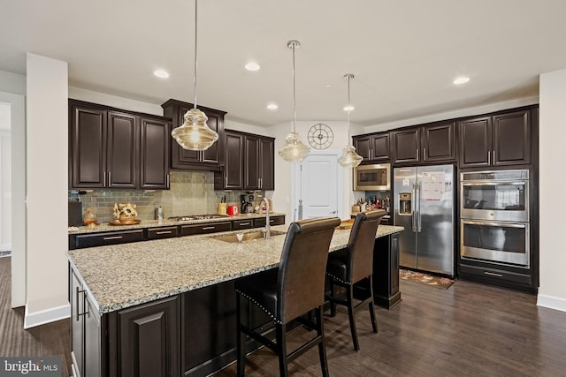 kitchen with dark hardwood / wood-style floors, decorative light fixtures, dark brown cabinets, a kitchen island with sink, and stainless steel appliances