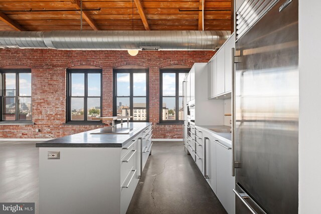 kitchen with white cabinetry, stainless steel built in fridge, brick wall, and a center island with sink