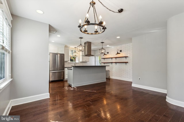 kitchen with wall chimney range hood, dark hardwood / wood-style flooring, hanging light fixtures, white cabinets, and stainless steel refrigerator