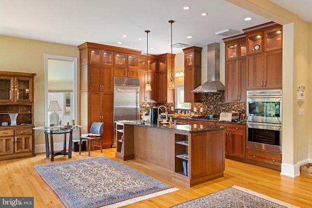 kitchen featuring decorative light fixtures, an island with sink, stainless steel appliances, dark stone countertops, and wall chimney range hood