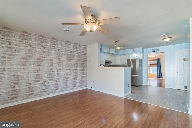 unfurnished living room with wood finished floors, wallpapered walls, baseboards, ceiling fan, and an accent wall