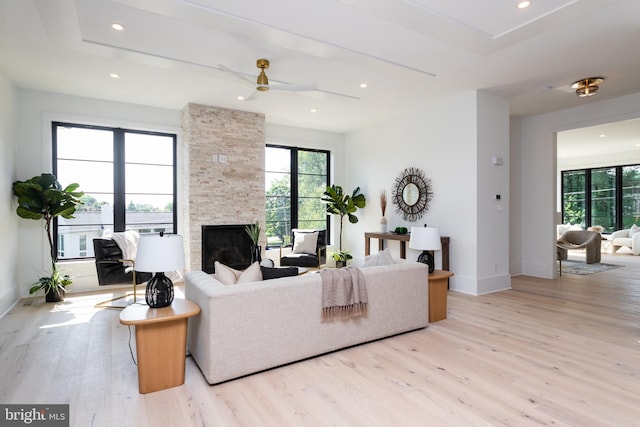 living room with ceiling fan, a stone fireplace, a tray ceiling, and light hardwood / wood-style floors