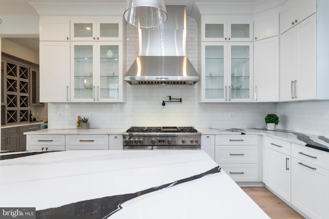 kitchen with wall chimney exhaust hood, light stone counters, white cabinets, and stainless steel range oven