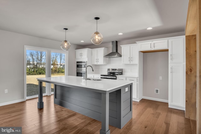 kitchen featuring a sink, wall chimney range hood, appliances with stainless steel finishes, white cabinets, and a kitchen island with sink