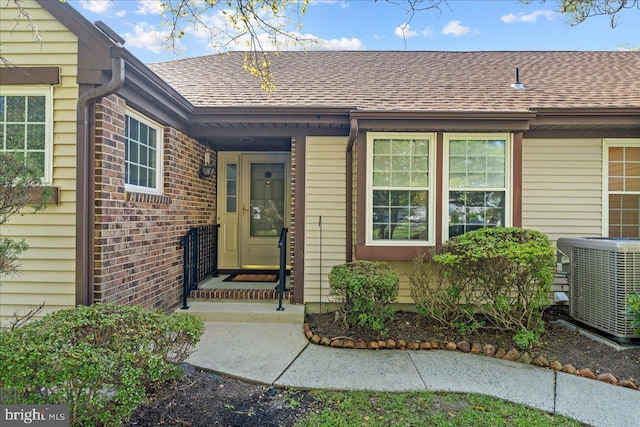 doorway to property featuring brick siding, central AC unit, and a shingled roof