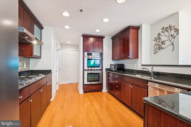 kitchen featuring exhaust hood, dark stone counters, sink, appliances with stainless steel finishes, and light hardwood / wood-style floors