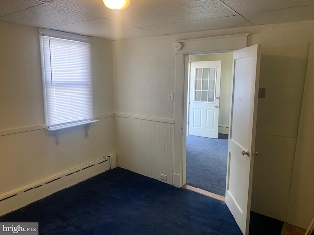 unfurnished room with a drop ceiling, a baseboard radiator, a wainscoted wall, and dark carpet