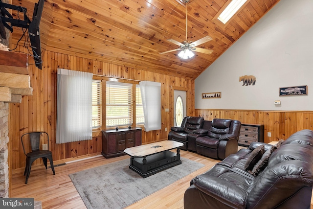 living room featuring wood walls, light hardwood / wood-style floors, high vaulted ceiling, a skylight, and ceiling fan