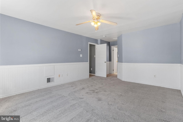 carpeted empty room with ceiling fan, visible vents, and a wainscoted wall